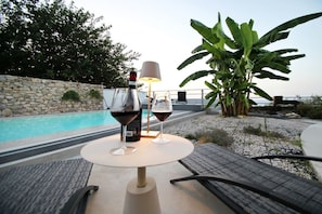Mesa com duas taças de vinho e uma garrafa, uma luminária, vista para uma piscina e vegetação.