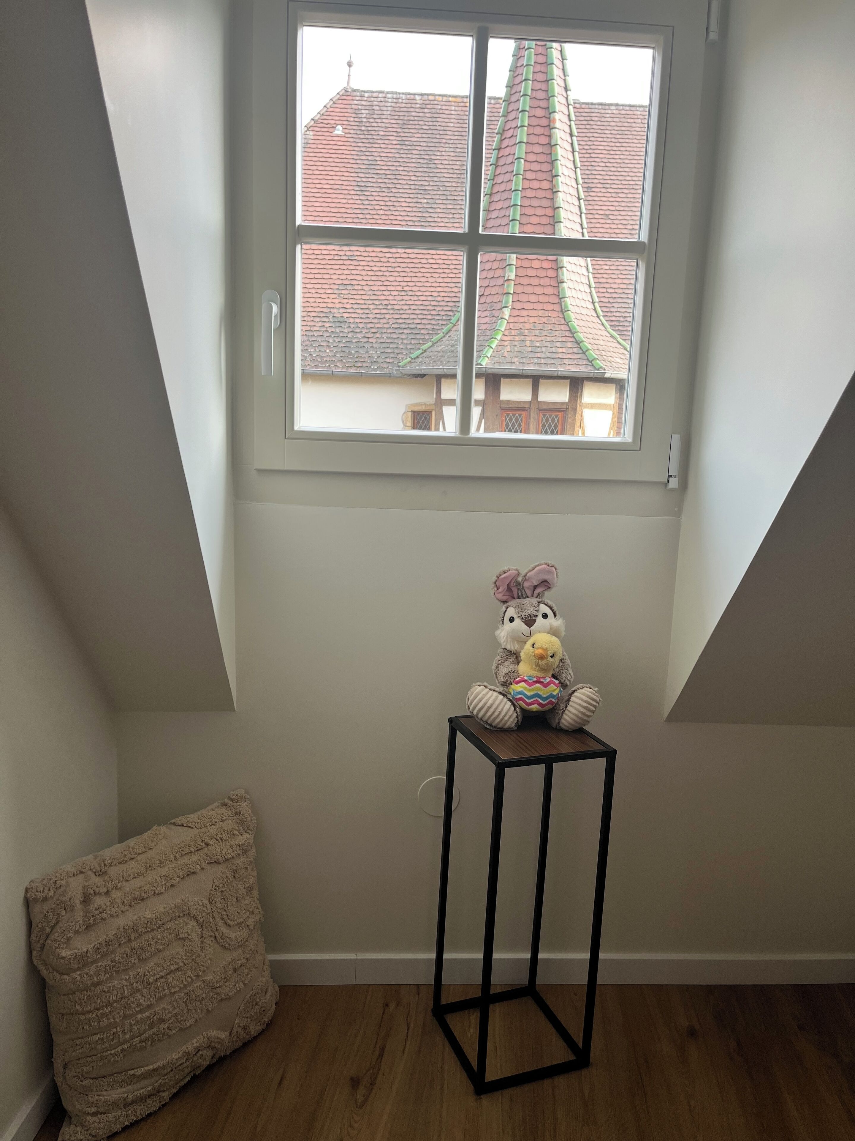 Ein Zimmer mit schrägen Wänden, einem Fenster mit Blick auf Gebäude, einem kleinen schwarzen Tisch und einem beigen Kissen.
