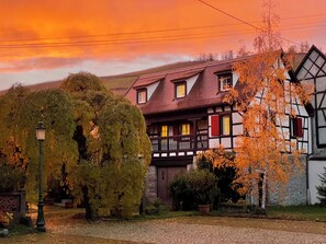 Ein traditionelles Fachwerkhäuschen mit Balkon, umgeben von Bäumen und einem Kopfsteinpflasterweg.