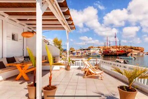 Eine Terrasse mit Blick auf einen Hafen, mit einem weißen Gebäude, Kübelpflanzen und Gartenmöbeln.