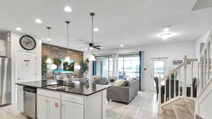 A modern kitchen with a central island, white cabinetry, and stainless steel appliances. There is a dining area with a gray sofa and a staircase leading to another level. - Lakefront 5BR Villa • Private Pool near Disney (Kissimmee)