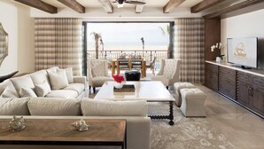 Een ruime woonkamer met een grote bank, een bijzettafeltje en een televisie op een kast. Er is een balkon met uitzicht op het strand.