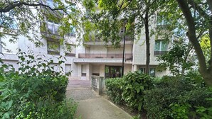 Ein Weg führt zu einem Gebäude mit einem eingenetzten Eingang, umgeben von Grünflächen und Bäumen.