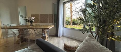 A modern living room with a wooden dining table, clear plastic chairs, a grey sofa, and a large window with a view of trees and a garden.