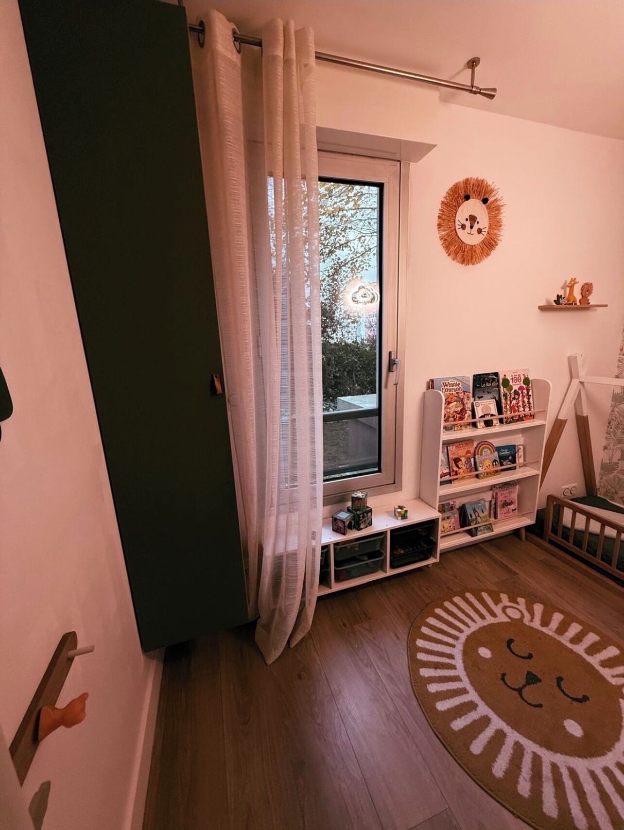 Una habitación infantil con ventana, estantería, alfombra con tema de león y decoración en la pared.