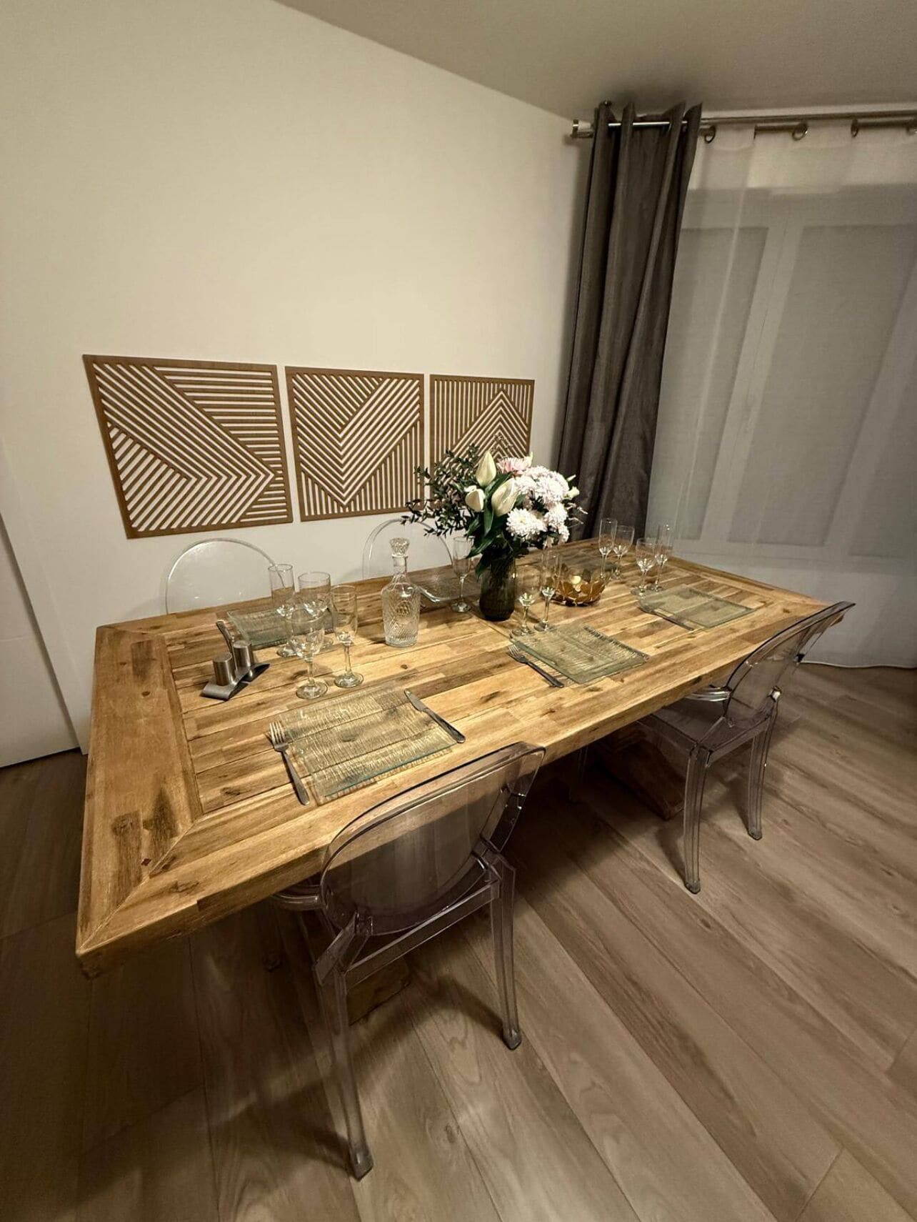 Ein Esstisch mit klarem Glas und einer Blumenvase, umgeben von klaren Stühlen in einem Raum mit Holzboden und dekorativen Wandverkleidungen.