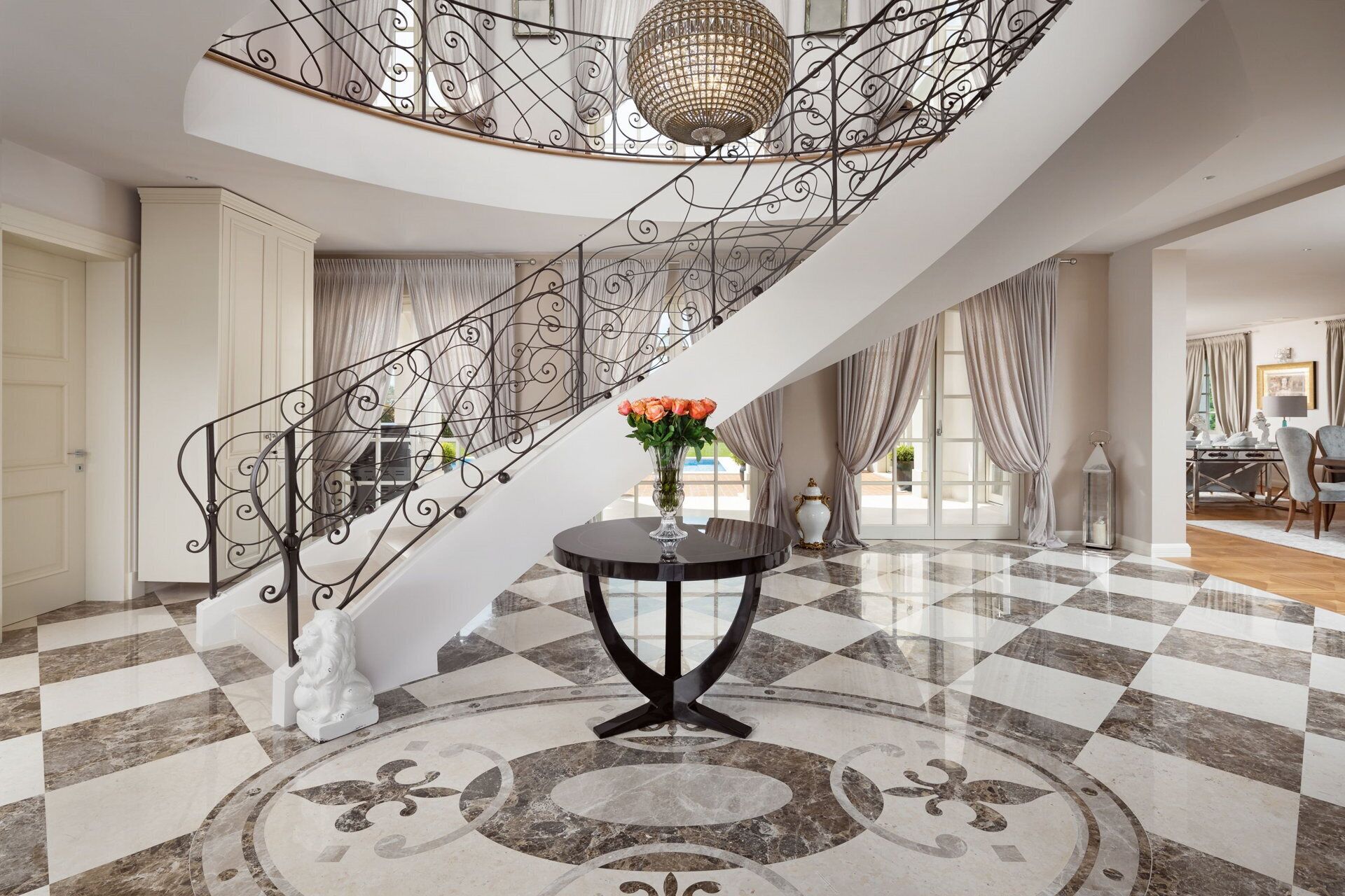 Un magnifique escalier orné d’une rampe en métal décorative et surmonté d’un lustre en cristal, menant à une pièce dont le sol est recouvert de carreaux de marbre et qui contient une table ronde agrémentée d’un vase de fleurs.