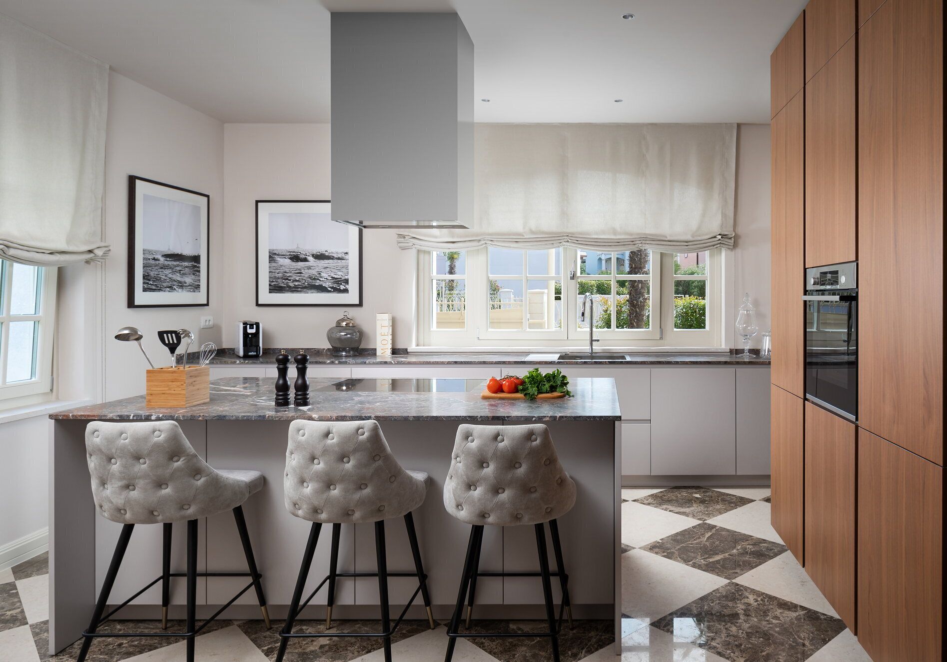 A modern kitchen with a large island, bar stools, a built-in oven, and a view of the outdoors through large windows.