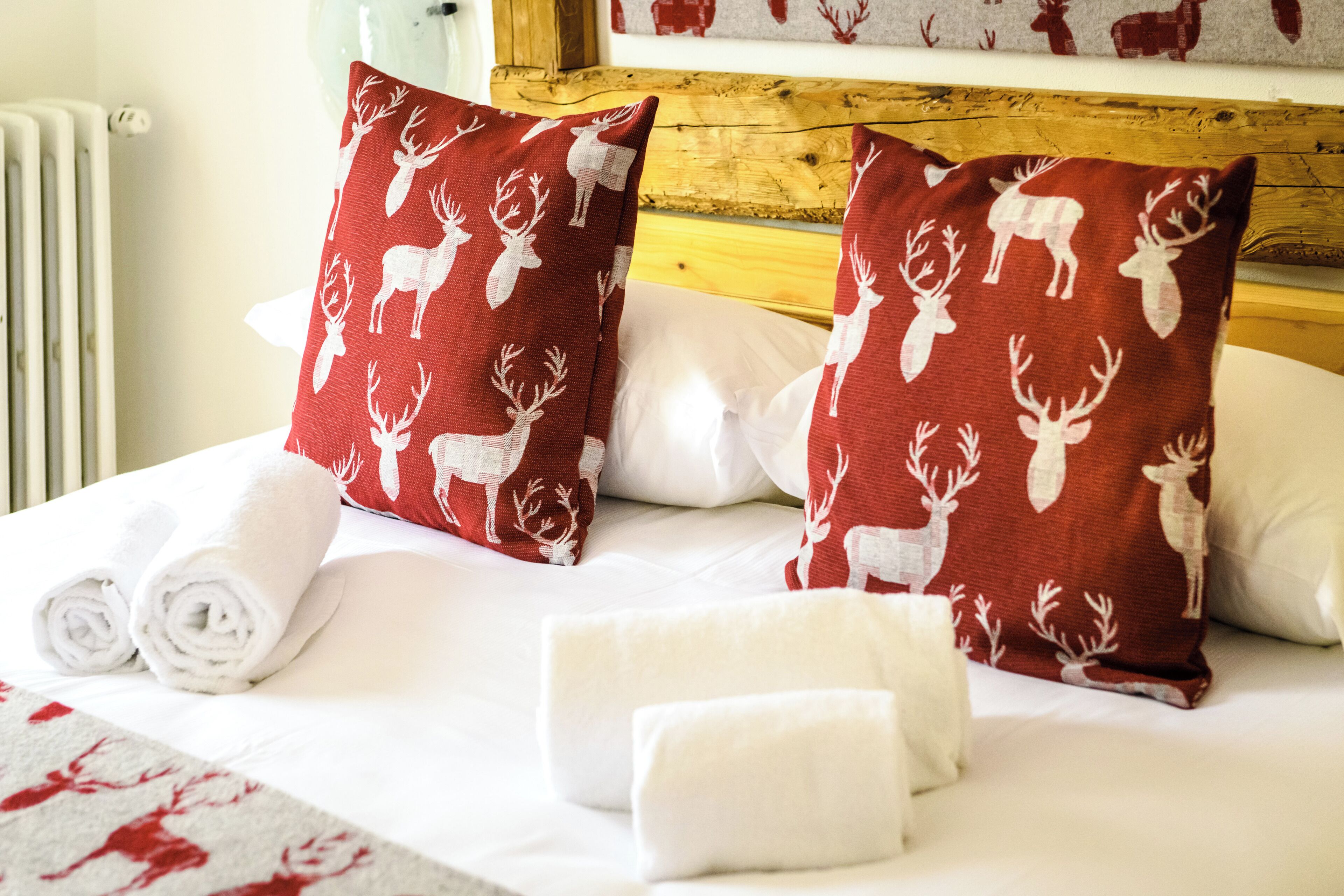 Una cama con ropa de cama blanca, dos almohadas rojas con estampados de ciervos blancos y toallas enrolladas de color blanco.