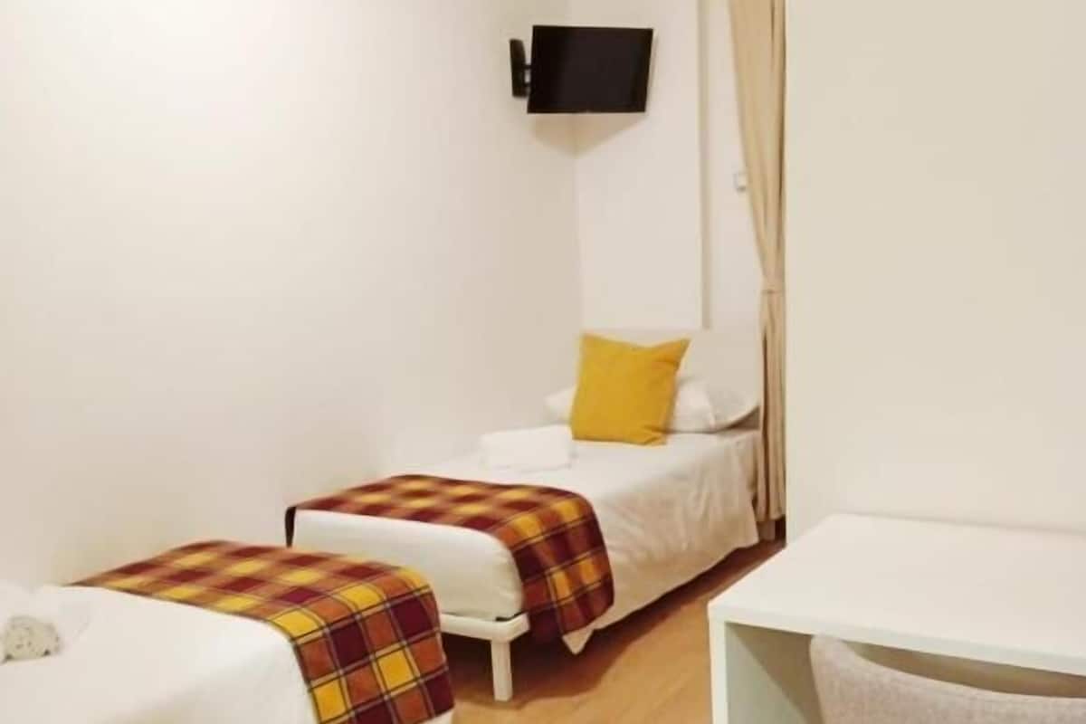 A small, compact room with two single beds, a small table, and a TV mounted on the wall.