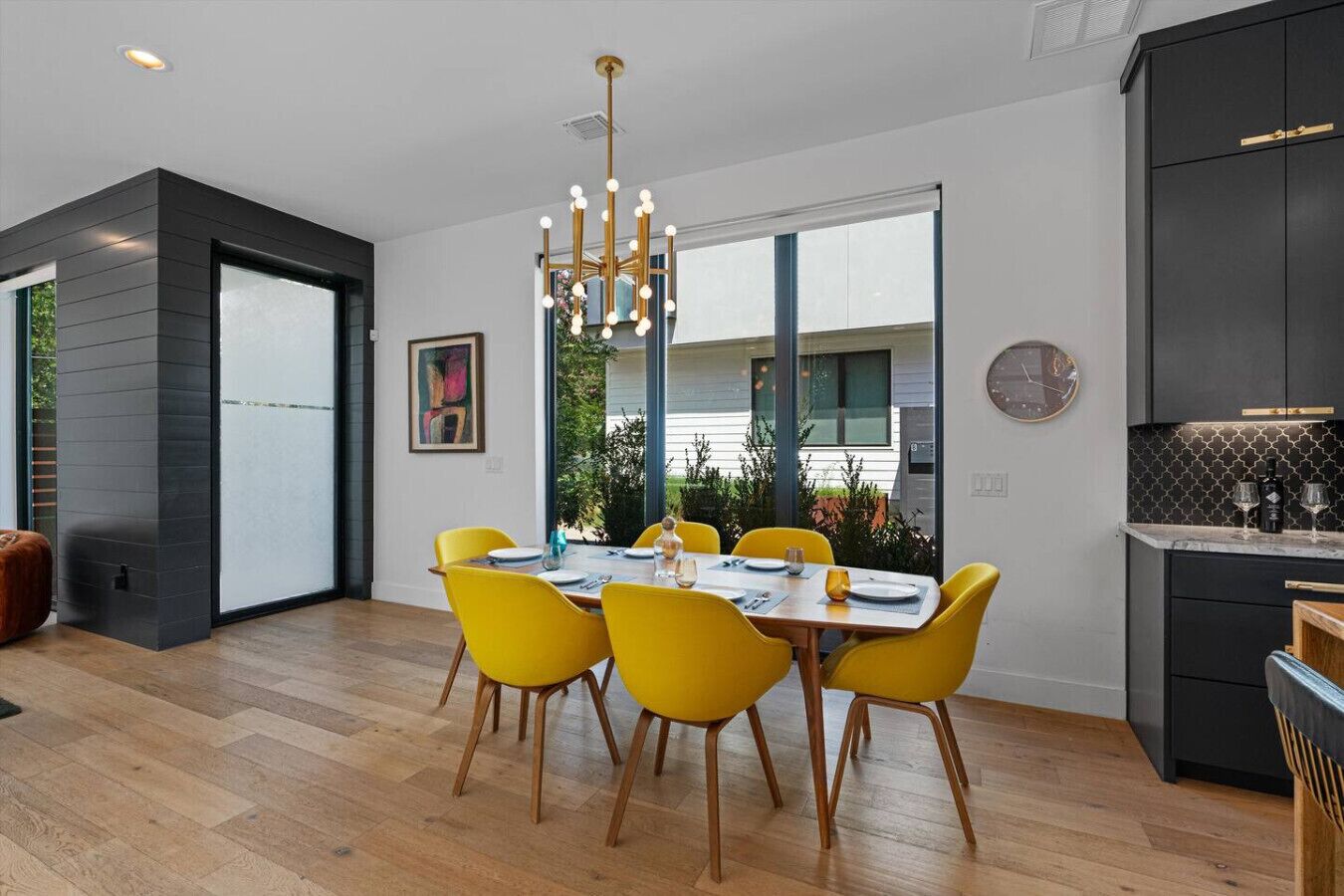 A modern kitchen with dark cabinetry, a dining area with yellow chairs, and a large window offering a view of greenery.