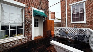 Un edificio de ladrillo con una puerta de madera, una puerta blanca y una ventana con marco blanco. Hay una mesa con sillas y una planta en maceta.