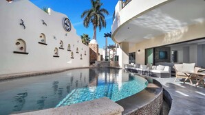 A modern pool area with clear water, a stone wall with decorative elements, and outdoor seating. - @ Marbella Lane – Grand Private Luxury Home Villa (Cabo Bello)