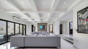 A spacious living area with a large sectional sofa, a stone accent wall, and a large window offering a view of the outdoors. - @ Marbella Lane – Grand Private Luxury Home Villa (Cabo Bello)