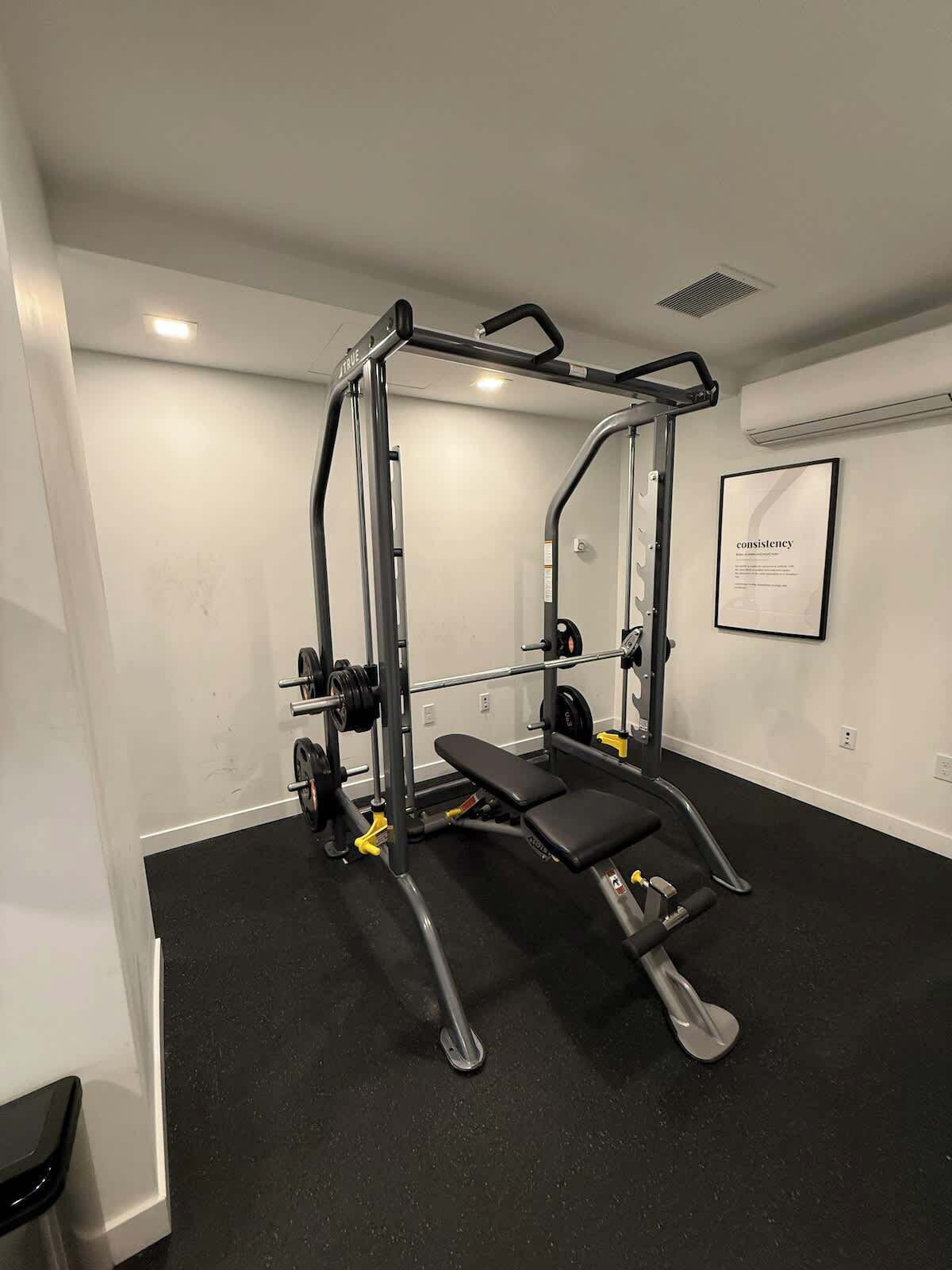 Un gimnasio pequeño con un soporte para sentadillas, banco y discos de pesas.