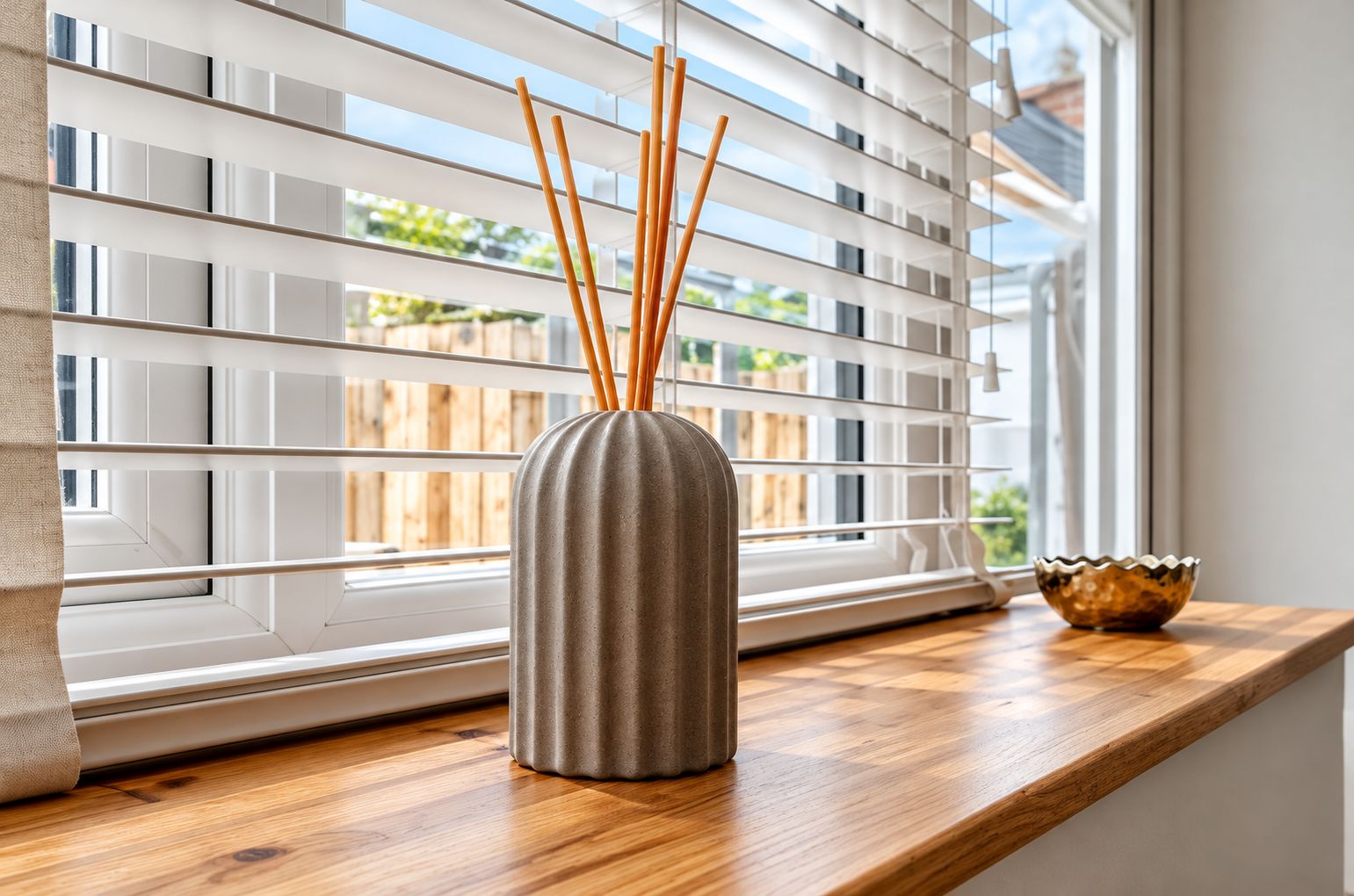 A window with white blinds, a vase with reed sticks, and a decorative bowl on a wooden ledge.
