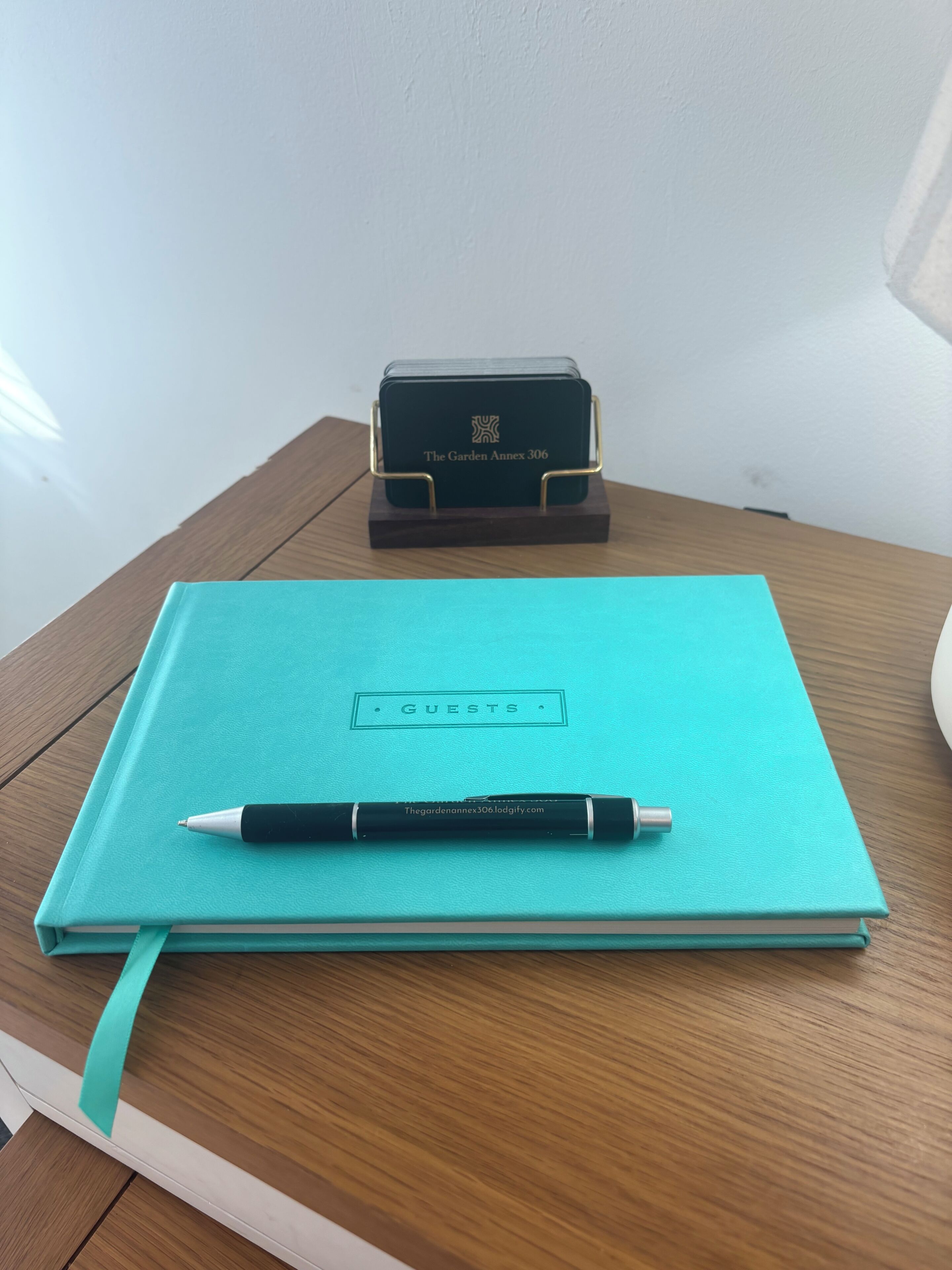 A teal notebook with a pen on top, labeled 'GUESTS,' and a black box with 'The Garden Annex 306' on a wooden table.
