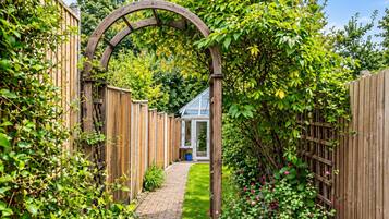 Un sendero en un jardín que lleva a una casa con un arco de madera, rodeada de abundante vegetación y una cerca de madera.