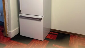 A white refrigerator in a corner of a room with a wooden floor and a brick wall.