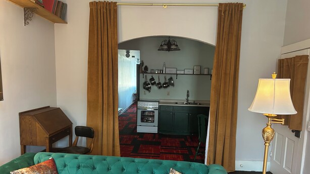 A living room with a green sofa, two decorative pillows, a lamp, and a view into a kitchen with a red patterned rug.