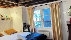 Une chambre d’hôtel avec un lit, une table de chevet, une fenêtre avec des rideaux, une étagère avec des livres et une vue sur les bâtiments environnants.