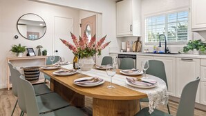 Un comedor con una mesa de madera dispuesta para cuatro personas, rodeado de sillas verdes. En la mesa hay un jarrón con flores rosas, y a la derecha una ventana con persianas.
