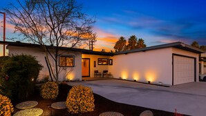 Una casa moderna con garaje, patio cubierto y jardín ajardinado con macetas de piedra y un árbol pequeño.
