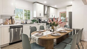 Una cocina moderna con armarios blancos, un lavavajillas de acero inoxidable y un comedor preparado para seis personas, con una mesa de madera y sillas grises.