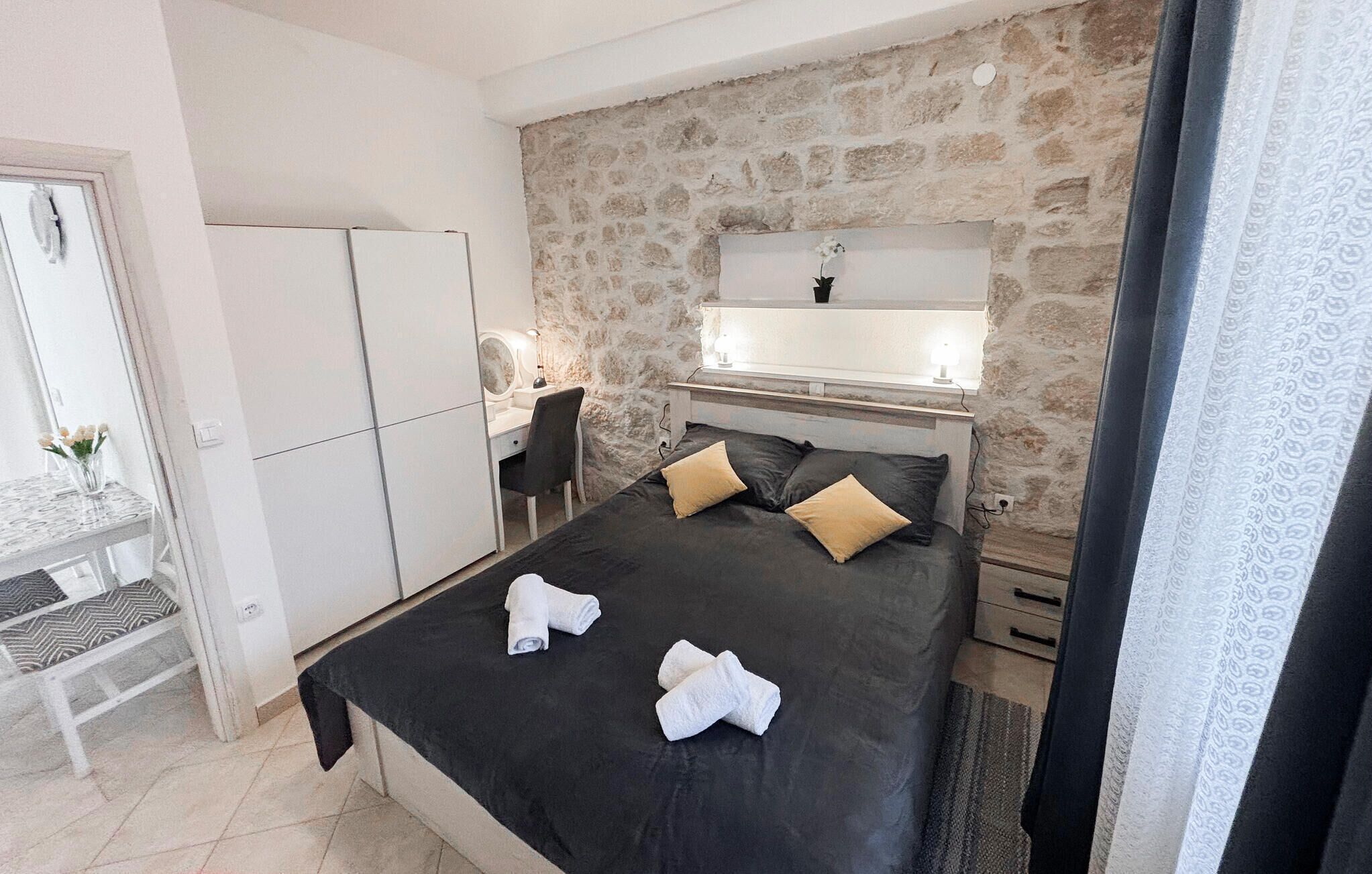 A bedroom with a stone wall, a bed with two pillows, a desk with a chair, and a mirror.