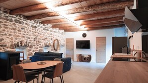 Un interior rústico con vigas de madera a la vista, una pared de piedra, una mesa de comedor de madera con sillas, un sofá y un televisor de pantalla plana montado en la pared.