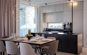 Un espace de repas moderne avec un îlot central, une cuisine en arrière-plan et une vue dégagée sur l’extérieur grâce à une grande fenêtre.