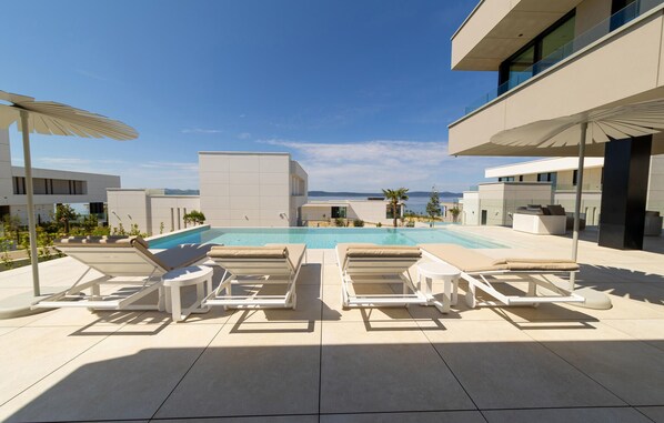 Un espace piscine extérieur moderne, doté de chaises longues et de parasols, entouré de bâtiments contemporains et offrant une vue dégagée sur la mer.