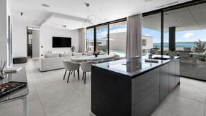A modern kitchen with a central island, a dining area with a round table and chairs, a living room with a sofa and a TV, and a view of the ocean through large windows. - Amazing home in Ugljan with private swimming pool, can be inside or outside (Ugljan)