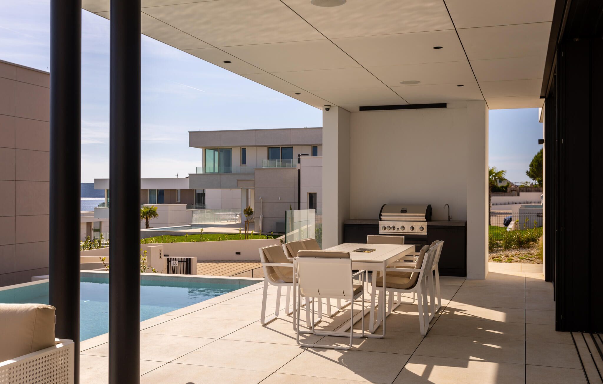 A modern outdoor patio with a pool, a barbecue grill, and a seating area with white chairs and a table.