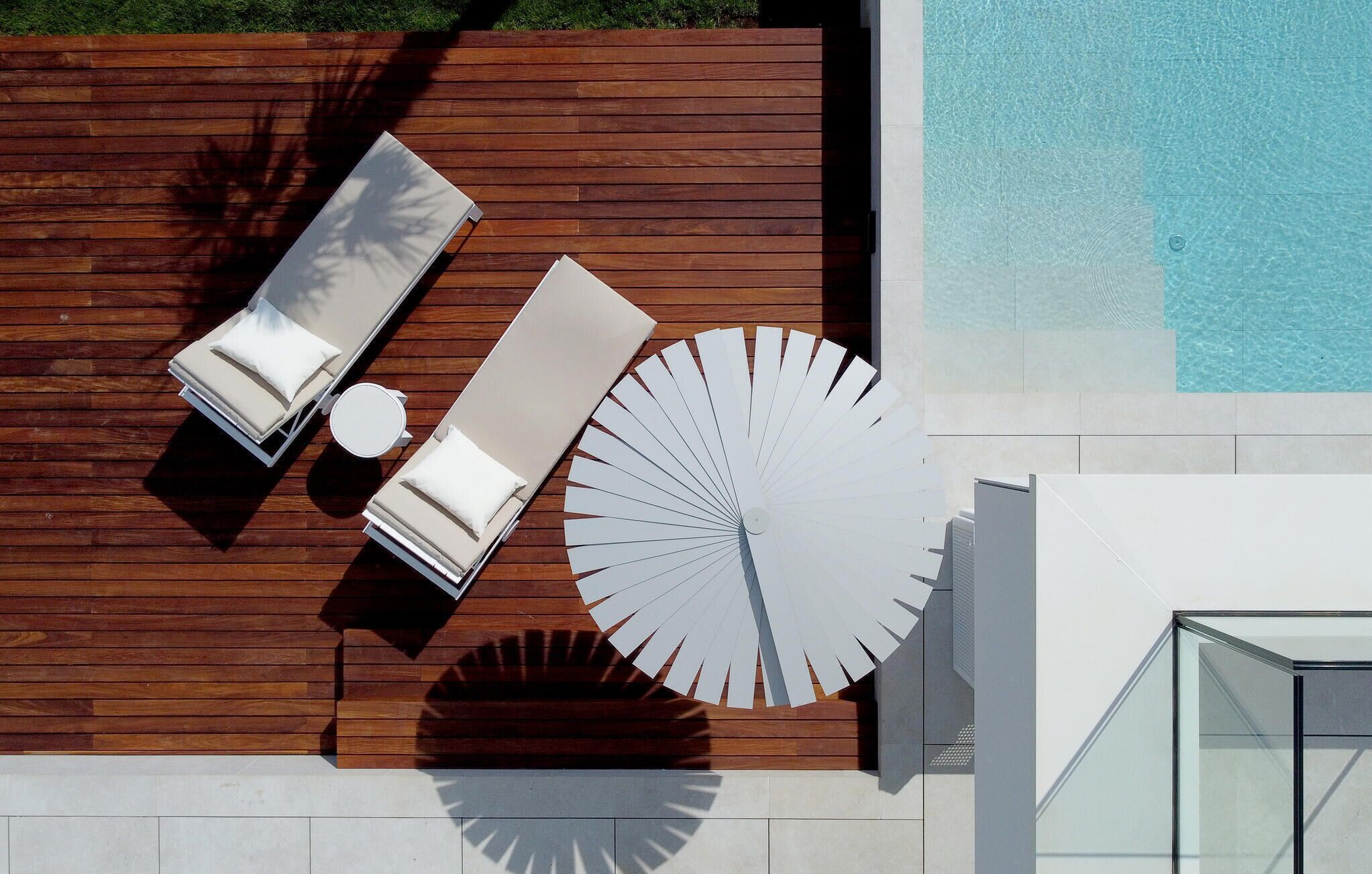 A rooftop pool area with wooden decking, two lounge chairs, a sun lounger, a large white fan, and a glass-enclosed structure.