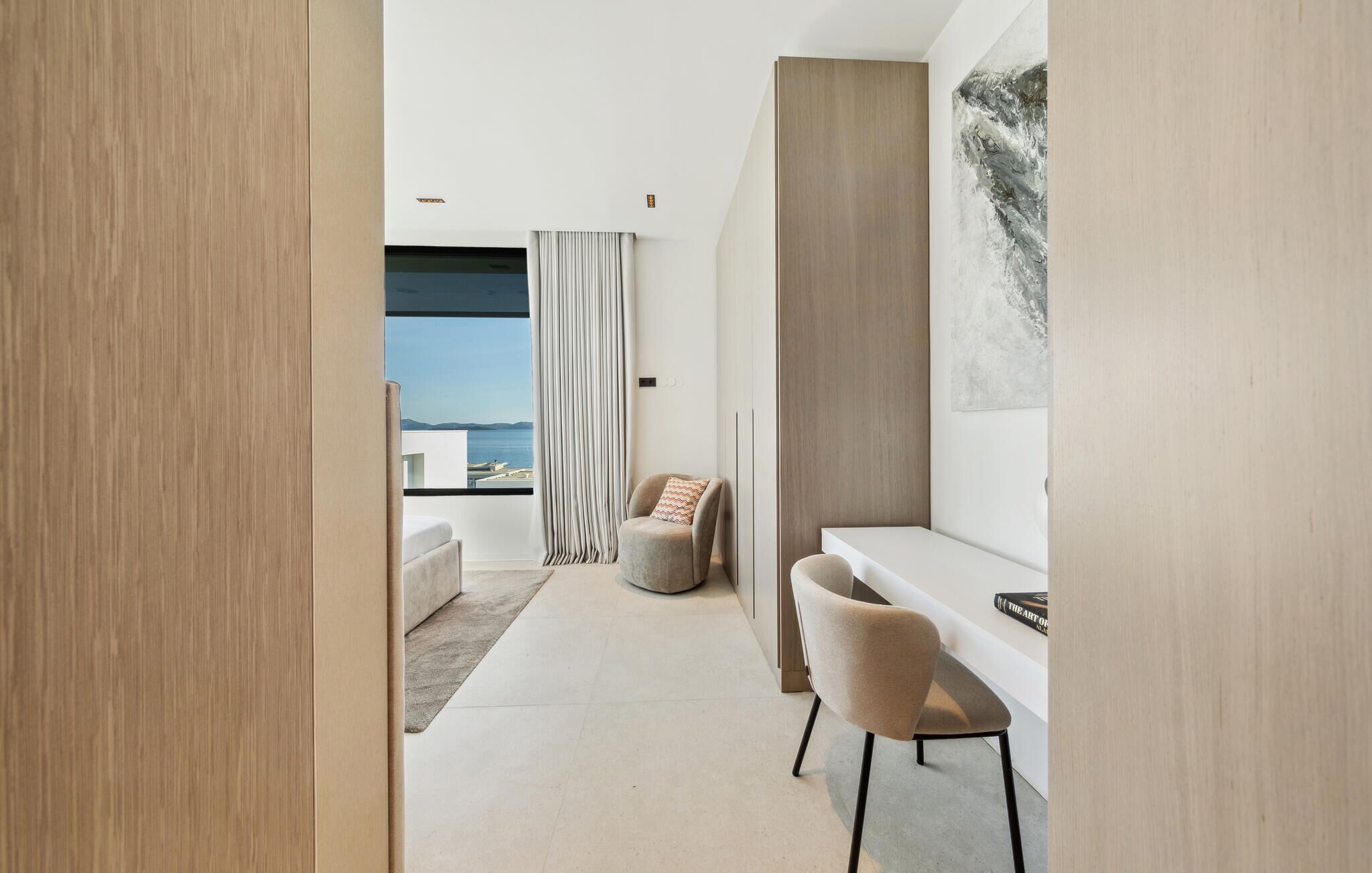 A modern hotel room with a minimalist design, featuring a large window with a view of the sea, a cozy seating area with a sofa and chairs, and a small desk with a chair.