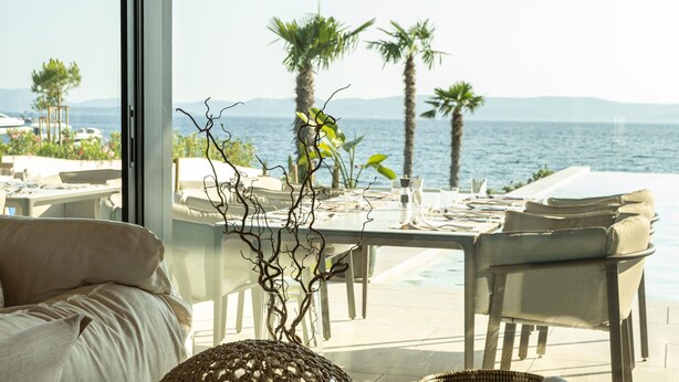 Un coin repas avec une table dressée pour deux personnes, offrant une vue sur la côte, avec des palmiers et la mer.