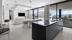 Eine moderne Küche mit einer zentralen Kochinsel, ein Essbereich mit rundem Tisch und Stühlen, ein Wohnzimmer mit Sofa und Fernseher sowie Meerblick durch große Fenster.