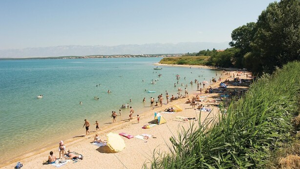Ein Strand mit klarem Wasser, Menschen beim Schwimmen und Sonnenbaden, und eine entfernte Bergkette im Hintergrund.