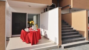 Un espace de restauration en plein air avec une table dressée pour deux personnes, recouverte d’une nappe rouge, et un vase rempli de fleurs jaunes.