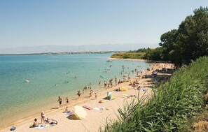 Ein Strand mit klarem Wasser, Menschen beim Schwimmen und Sonnenbaden, und eine entfernte Bergkette im Hintergrund.