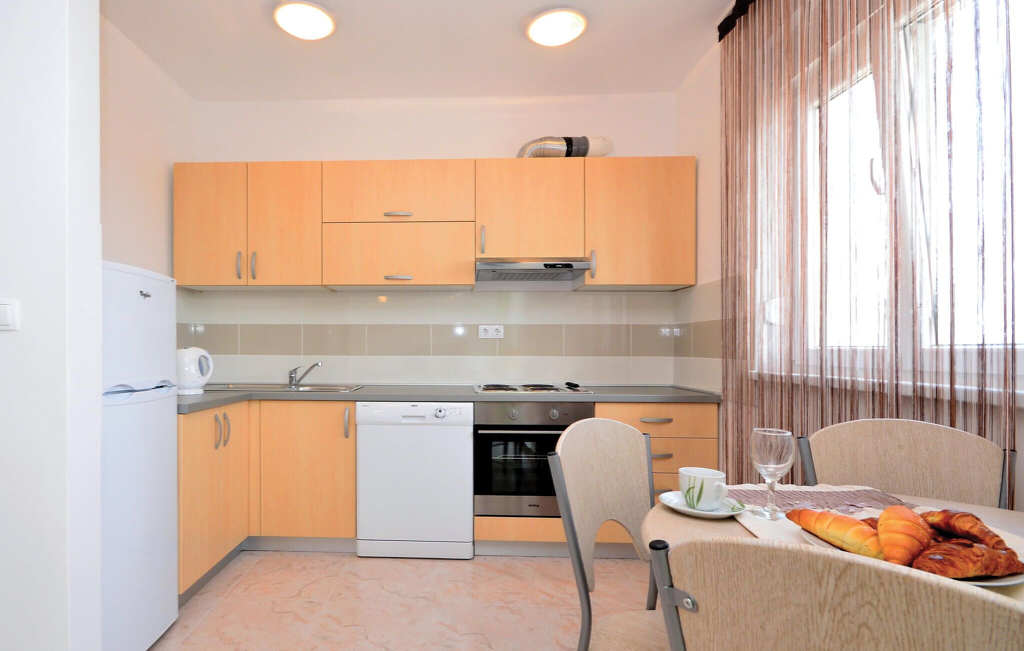 A compact kitchen with a dishwasher, stove, and oven. There is a dining area with a table set for two, including a croissant and a glass of water.