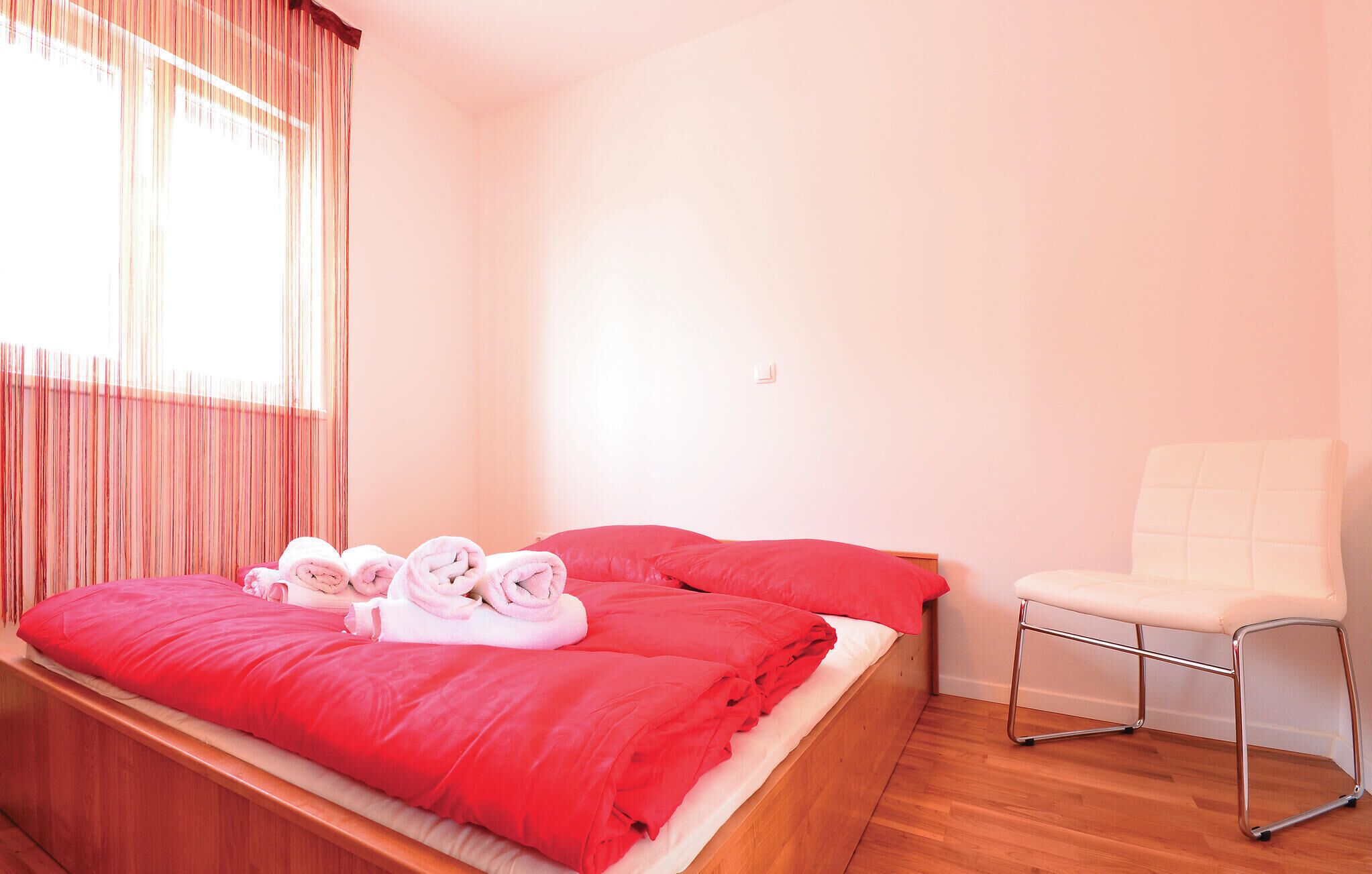 Ein Schlafzimmer mit einem hölzernen Bett, roter Bettwäsche, einem weißen Stuhl und einem Fenster mit Vorhängen.