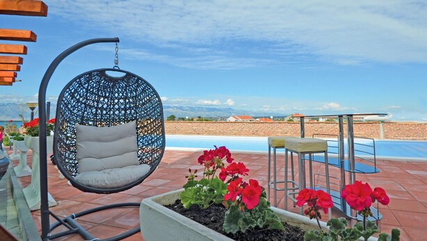 Una silla colgante con cojín, una piscina y flores rojas en una maceta.