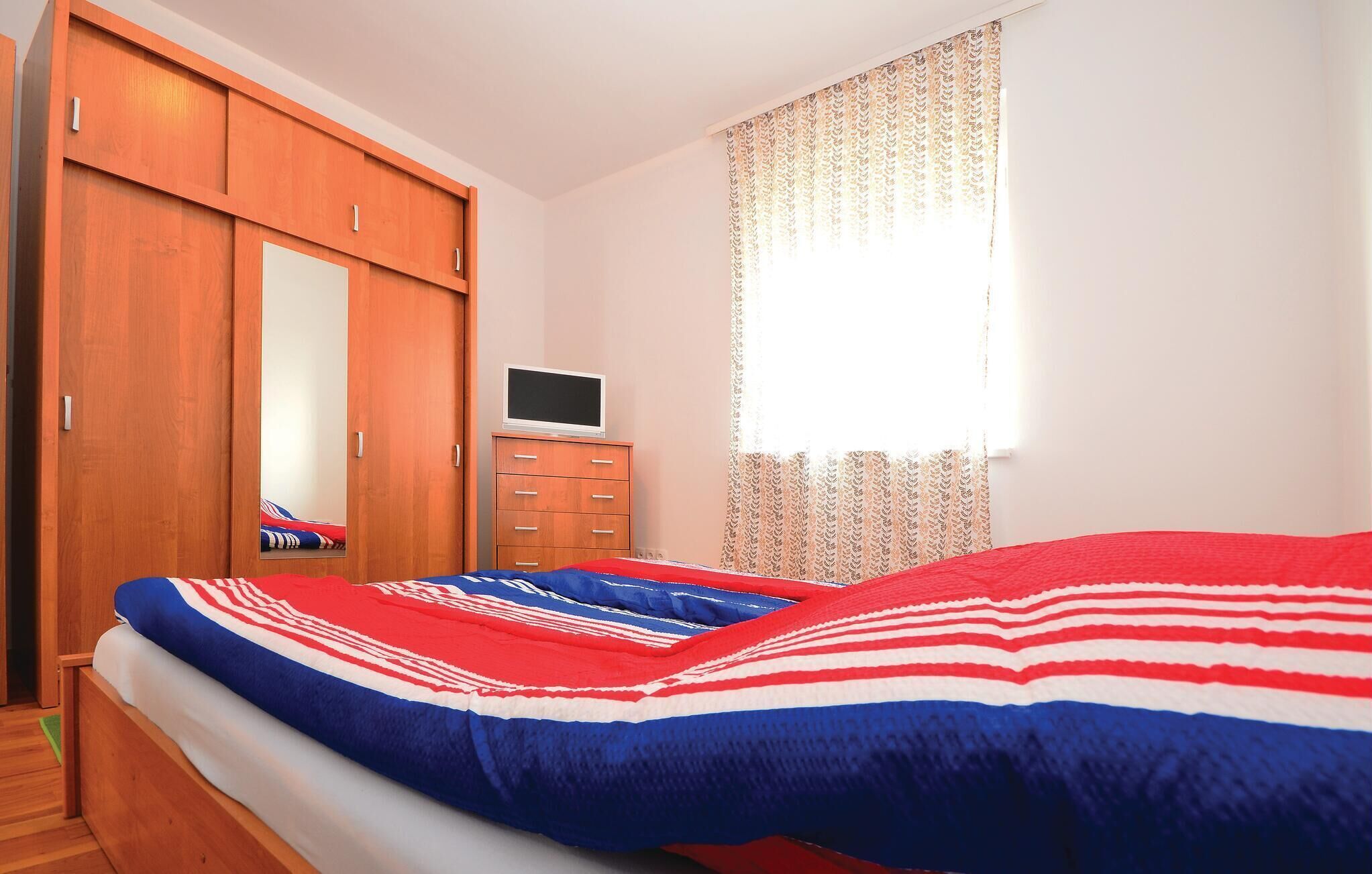 Un dormitorio con cama, armario, televisor sobre un aparador y ventana con cortinas estampadas.