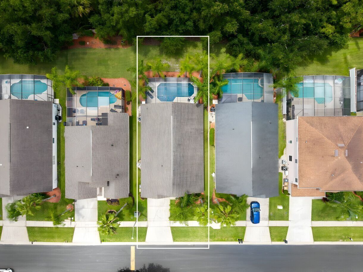 An aerial view of a residential area with multiple houses, each with a swimming pool, surrounded by greenery and a road in the foreground.