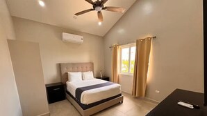Une chambre avec un lit, un ventilateur de plafond, un appareil de climatisation, une fenêtre avec des rideaux et un bureau avec une télécommande.