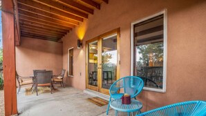 A covered patio with a table and chairs, a large window, and a door leading to an interior space. - Cielo Grande · Near Santa Fe Plaza, Small Fenced Yard, Parking (Santa Fe)