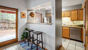 A compact kitchen with wooden cabinets, a stainless steel refrigerator, and a built-in oven. There are two stools near the entrance and a small window with a view of the outside. - Cielo Grande · Near Santa Fe Plaza, Small Fenced Yard, Parking (Santa Fe)