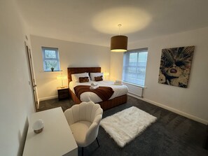 Een moderne slaapkamer met een groot bed, nachtlampjes, een witte stoel en een zacht vloerkleed.